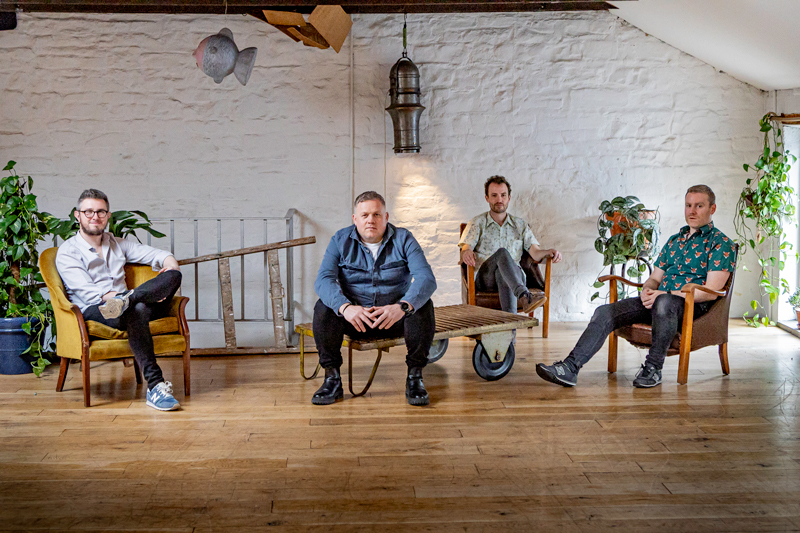Aldoc band photo. The four musicians sitting in a spacious room. Three sit on chairs and one on a push cart.
