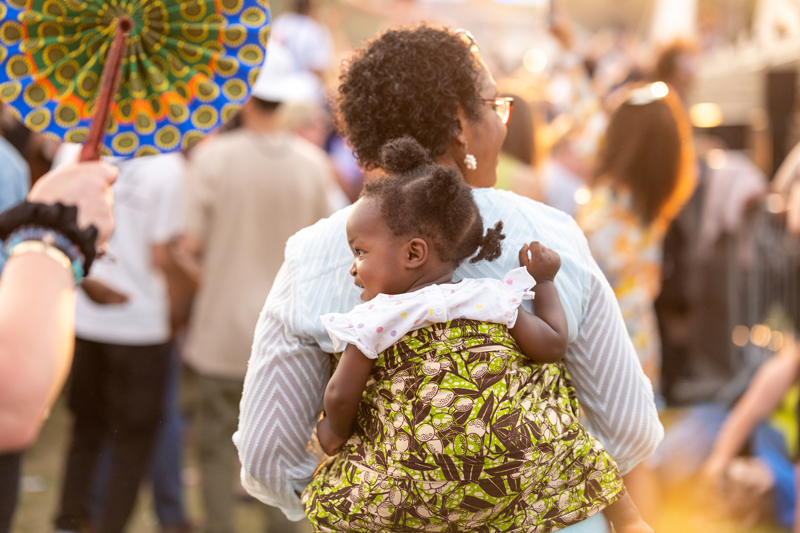 Africa Oyé 2023 - Photo Mark McNulty. a photo of festivalgoers with a black woman carrying her daughter on her back.