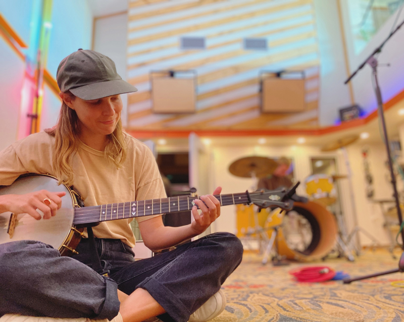 Allison de Groot - Photo by Phil Cook. Allison sitting on the ground outdoors, playing banjo.