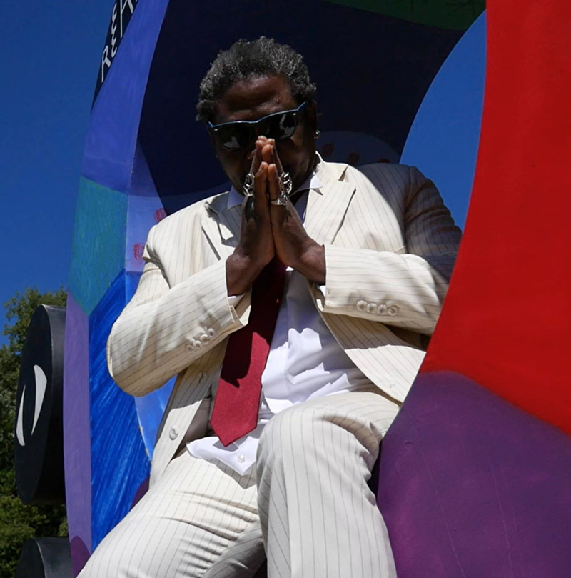 The single cover features Angelo Rascool in a cream pinstripe suit and red tie, seated with hands pressed in a prayer gesture. He's wearing dark sunglasses and rings. Set against a geometric backdrop under clear blue skies.