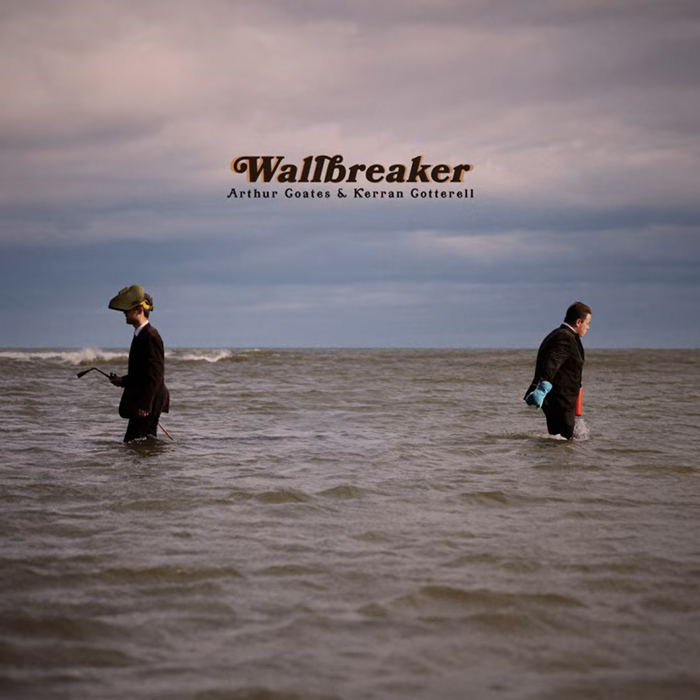 Two men in suits stand waist-deep in the sea, facing away from each other. One wears a strange helmet and holds a device, while the other carries bright cleaning gloves. Centered text above them reads “Wallbreaker” with the artists’ names beneath it.