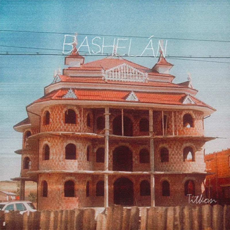 A photograph of an unfinished multi-story brick building with arched windows and a red-tiled roof under a blue sky crossed by power lines. Handwritten white text reading Bashel Án floats above the structure, while Titkon appears in the lower corner.