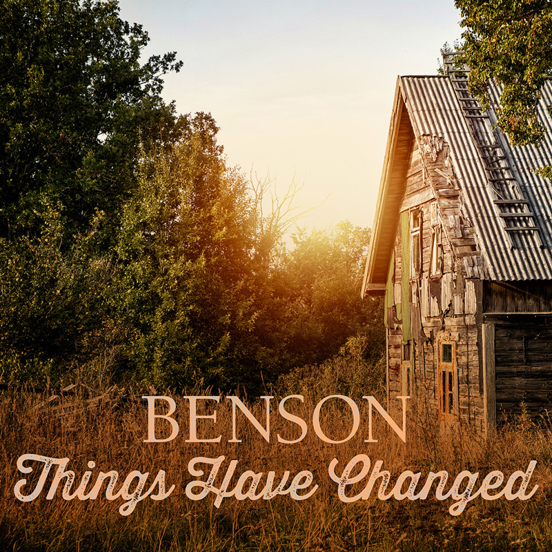 The single cover for Things Have Changed by Benson features a rustic, weathered wooden house with a metal roof, surrounded by overgrown grass and lush green trees. The setting is bathed in warm, golden sunlight.