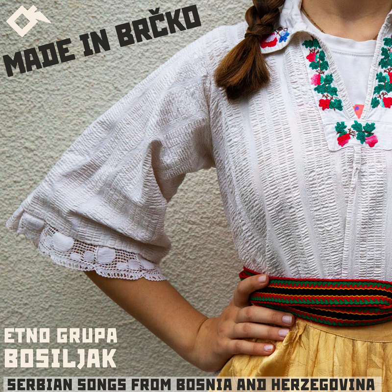 The album cover for Etno Grupa Bosiljak – Made in Brčko: Serbian Songs from Bosnia and Herzegovina features a close-up of a young woman in traditional folk attire. she wears an embroidered white blouse, colorful woven belt, and golden skirt.