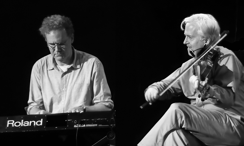 Brian Conway playing fiddle live accompanied by a keyboard player. Black and white photo.