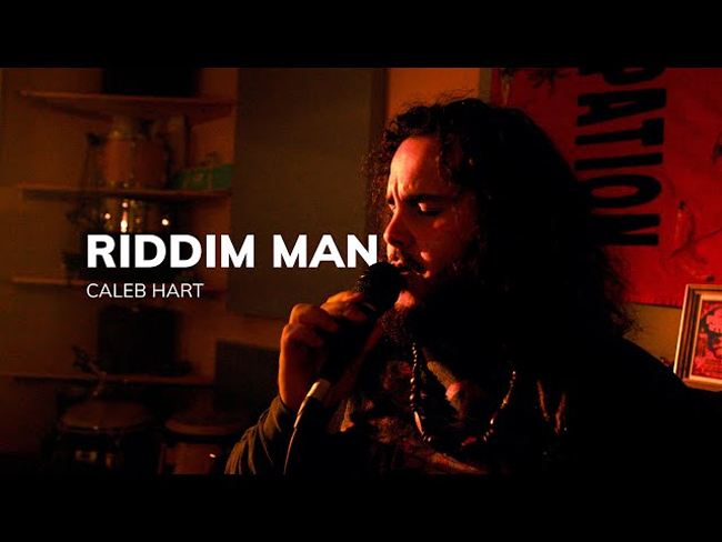 Caleb Hart Riddim Man video screenshot. The artist singing, holding a microphone.