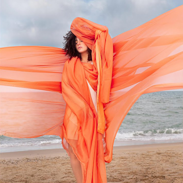 Carmel stands on a sandy shoreline beneath a pale sky, wrapped in sweeping, translucent orange fabric that lifts in the sea breeze.