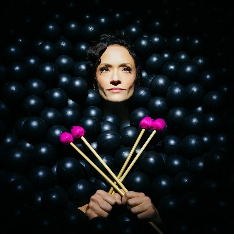 Dark spheres envelop Carolyn-Trowbridge. she holds bright pink mallets that cross in her hands.