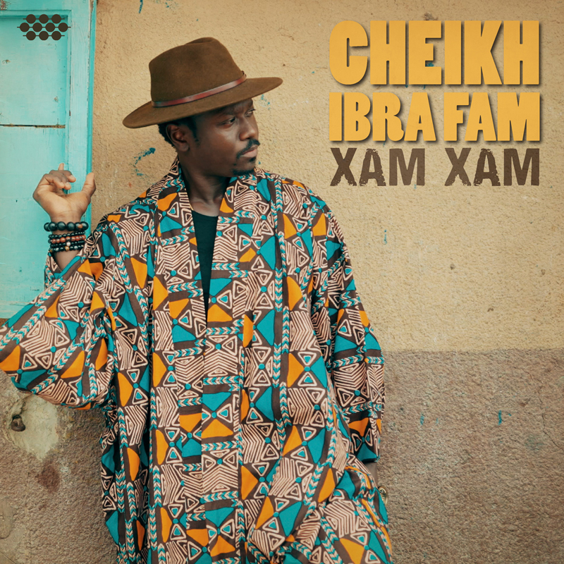 The single cover for "Xam Xam" by Cheikh Ibra Fam features the artist standing against a textured, two-toned wall—light tan on top, faded mauve below. Dressed in a geometric-patterned robe of teal, orange, black, and cream, he gazes to the side beneath a brown fedora. His left hand is slightly raised, adorned with beaded bracelets.