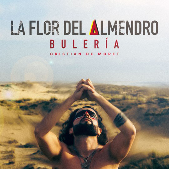 The cover of Cristian de Moret's single La Flor del Almendro (Bulería) features the artist in a desert-like landscape, arms raised in a posture of devotion or surrender. He wears sunglasses and a bare chest. Above him, the title appears in bold stencil-like typography, with the letter "A" stylized as a red triangle with a yellow accent.