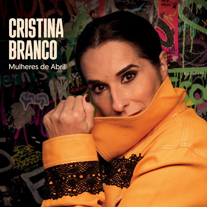 Cristina Branco in an orange jacket pulls the collar toward her face, meeting the camera. Behind her, a wall layered with graffiti. White lettering at the side announces the album title.