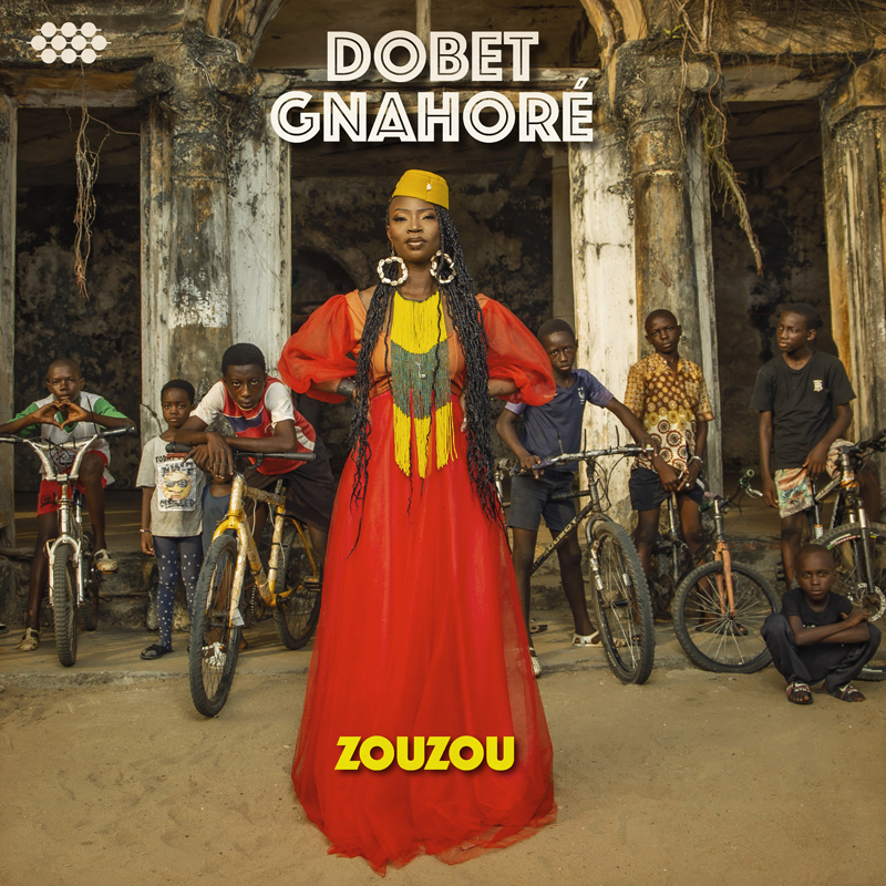 Dobet Gnahoré - Zouzou cover artwork. The artist standing in the street, wearing a red dress in front of several children with bicycles.