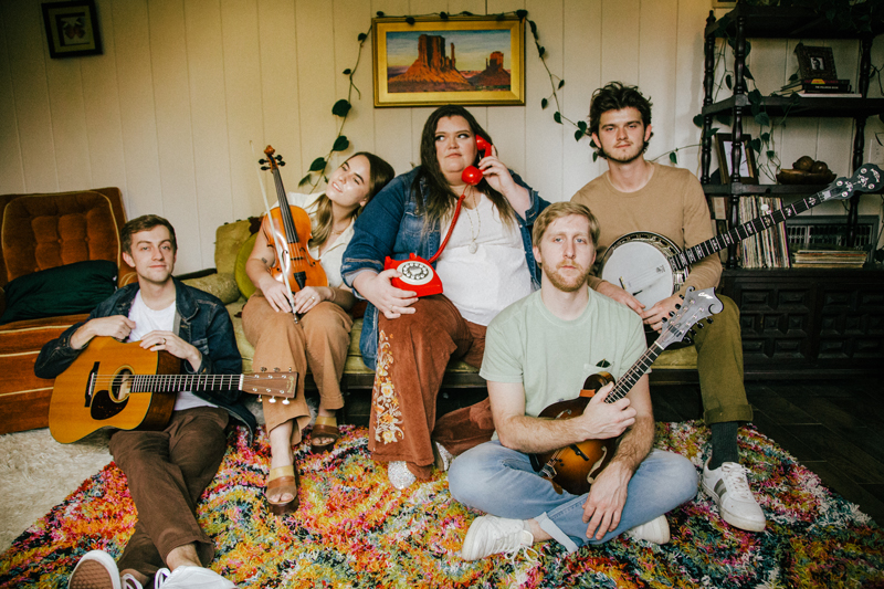 DownRiver Collective in 2023. The musicians sitting in a living room holding their musical instruments. One of them is holding an old style landline red phone.