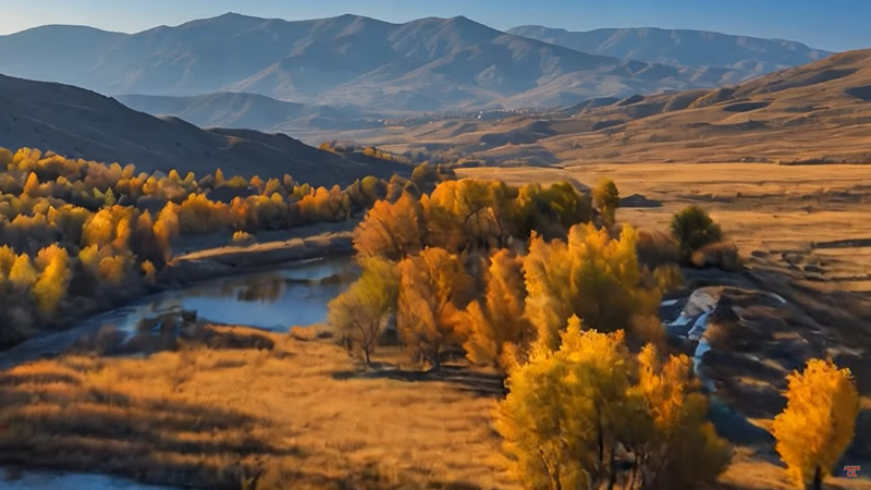 This image extracted from the AI generated video of the song Ela Gözlerini Sevdiği Dilber depicts a serene autumn landscape featuring a golden-hued valley with colorful trees surrounding a calm stream. Rolling hills and majestic mountains frame the background under a clear blue sky.