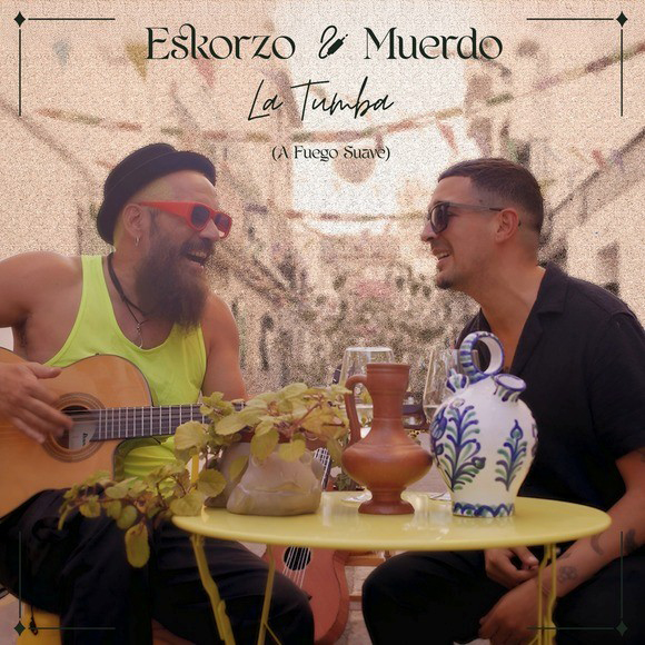 Eskorzo & Muerdo sit outdoors at a small round table, smiling at one another. One plays a guitar while wearing sunglasses and a hat, the other wears dark sunglasses and an open shirt. Ceramic jugs, wine glasses, and a plant decorate the table. The cover text introduces a collaboration titled La Tumba (A Fuego Suave) by Eskorzo & Muerdo.
