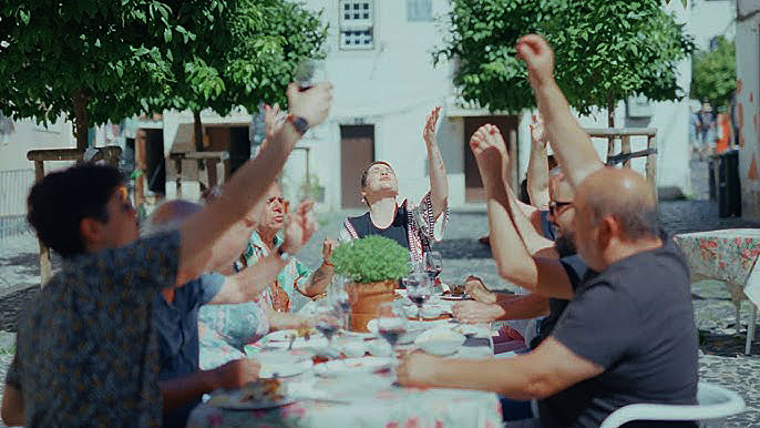 Fábia Rebordão has released her new single, “A Voar Por Cima Das Águas. a phot extracted from the video showing people sitting around a tabe outdoors eating and drinking.