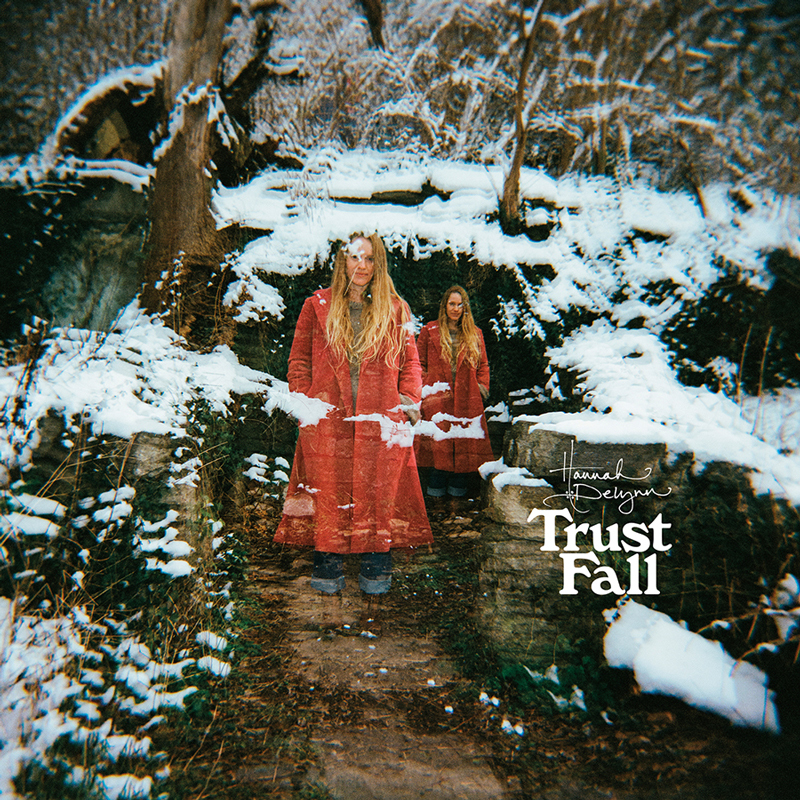 The album cover for Trust Fall by Hannah Delynn features a double-exposed photograph of a woman in a long red coat standing on a snow-dusted forest path. The blurred layering effect creates the illusion of her appearing twice, side by side, against a backdrop of bare trees and stone steps. The title Trust Fall and artist’s name appear in handwritten-style white lettering in the lower right corner.