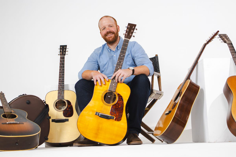 Jesse Smathers - Photo by Laci Mack. Jesse sitting on a chair, holding a guitar. Surrounded by various guitar models.