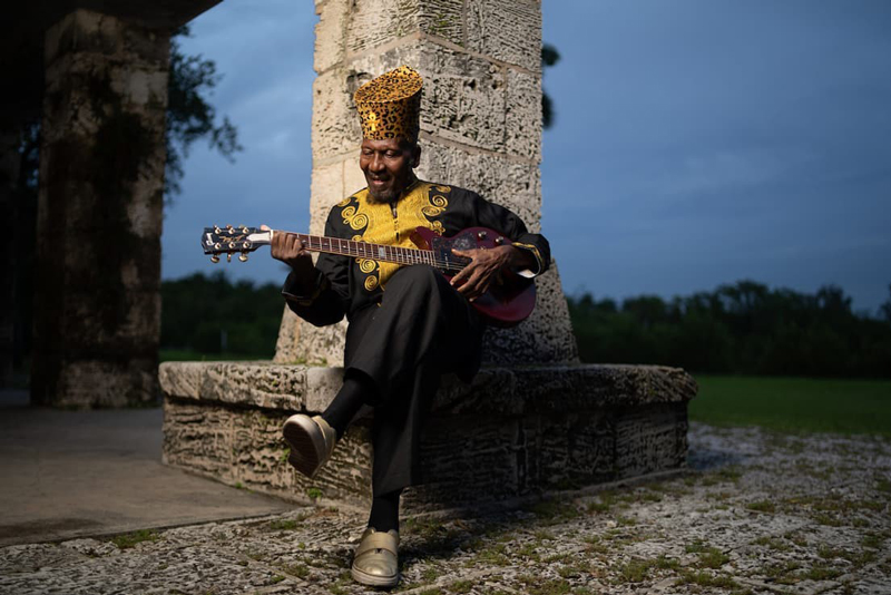 Jimmy Cliff - Photo credit Vision Addict. Jimmy Cliff, sitting outdoors, playing a guitar.