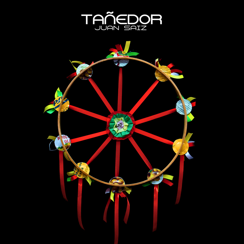 Juan Saiz - Tañedor cover artwork. A black album cover featuring a handmade circular wheel-like sculpture with red spokes, ribbons, and colorful recycled-material ornaments. The title “TAÑEDOR” and artist name “JUAN SAIZ” appear in white lettering at the top.