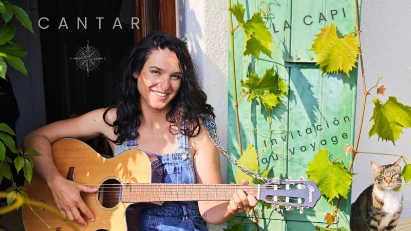 a photo of La Capi sitting in a garden, smiling, holding her guitar, next to a cat.