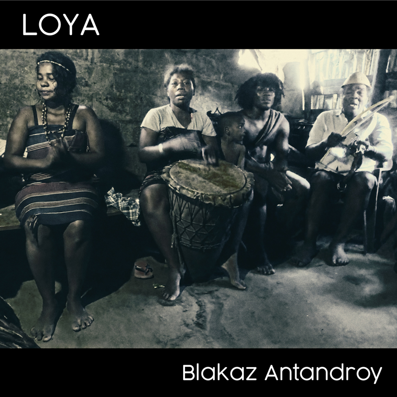 This album cover for Blakaz Antandroy by Loya features a monochromatic photograph with a vintage, earthy tone. It portrays a group of people seated in what appears to be a modest, rustic indoor setting. The central figure is playing a traditional drum, while others participate in the scene, possibly singing or clapping.