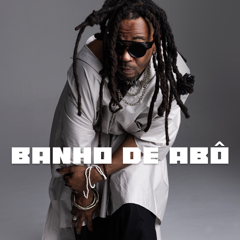 The single cover for Banho de Abô by Mamah Soares features the artist in a dynamic pose in a flowing white tunic, accessorized with chunky jewelry and dreadlocks.