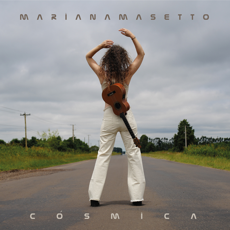 Mariana Masetto - Cósmica cover artwork. The artist standing in the middle of a road with a guitar on her back, wit her back to the camera.