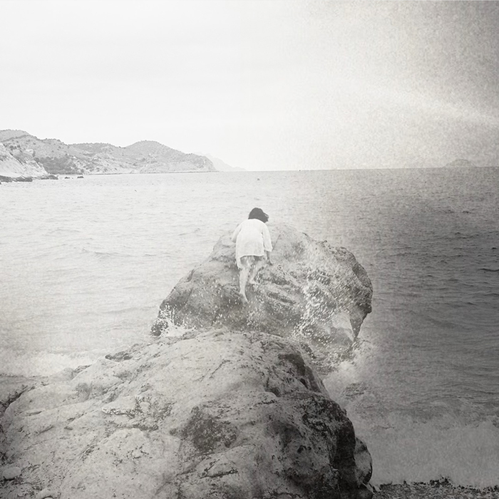 Marina Paredes - Para Nacer cover artwork. A woman standing at the edge of the shore with the sea and far away mountains behind.