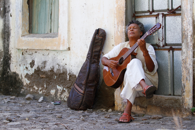 Mario Lucio - Photo by Omar Camilo. The artist sitting by a wall, playing guitar.