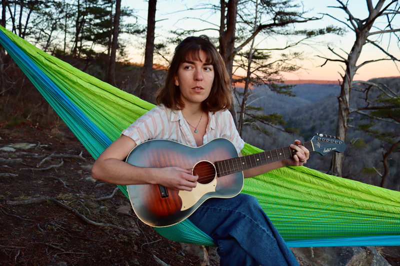 Maya de Vitry playing guitar sitting on a hammock. Photo by Ethan Jodziewicz.