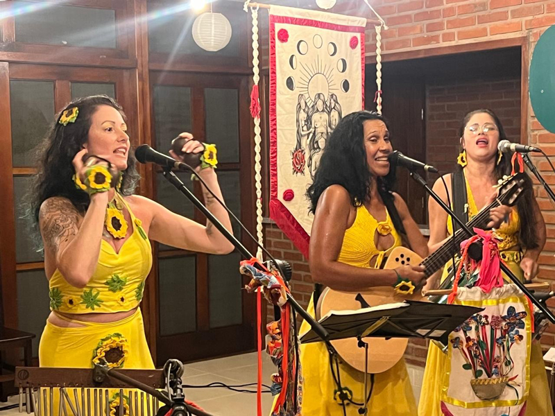 Meninas do Sol performing on stage. Three women in yellow dresses playing musical instruments.