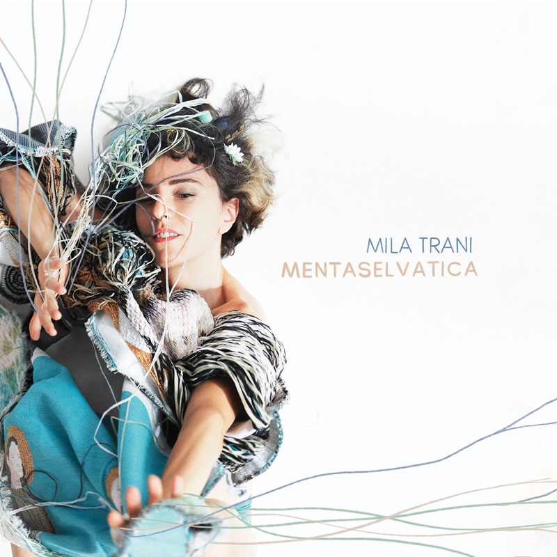 The album cover for Menta Selvatica by Mila Trani features a central figure, presumably the artist, draped in a textured, eclectic outfit with woven elements, soft fabrics, and intricate patterns. Her expression is serene yet expressive, with wild, tousled hair adorned with delicate details like thin wires and small flowers. The background is white.