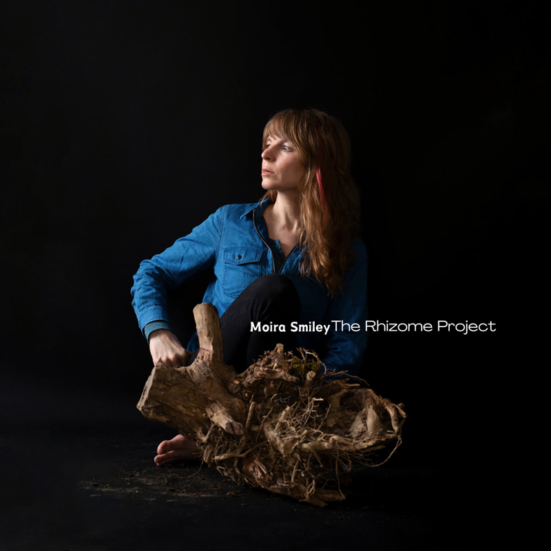 Moira Smiley – The Rhizome Project cover artwork. A photo of Moira sitting barefooted on the floor in front of a tree root and rhizome.