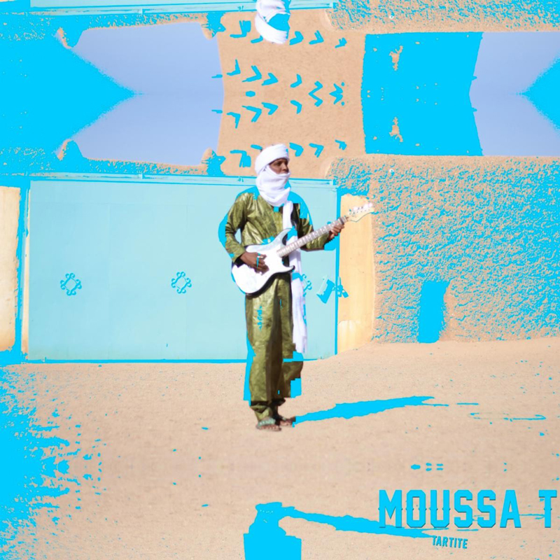 A desert scene with Moussa in traditional Tuareg-style clothing: flowing robe and face covering. He is standing barefoot in front of a wall painted in urquoise and sand tones. He holds a white electric guitar.