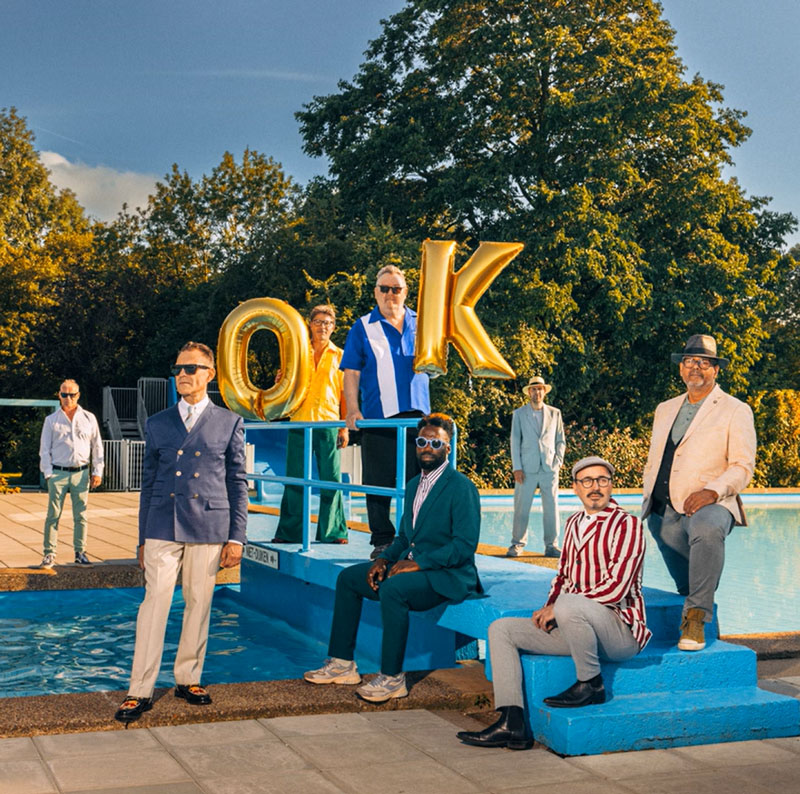 The New Cool Collective's Everything Is OK album cover features a group of stylishly dressed musicians posing around an outdoor swimming pool under a clear blue sky. The scene exudes a retro-cool aesthetic with bold colors, tailored suits, and vintage-inspired fashion. Two members hold large gold balloons forming the letters "O" and "K." The backdrop features lush green trees and warm lighting.