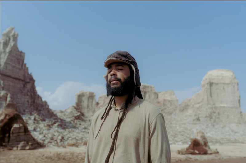 Protoje standing outdoors. Behind him, a landscape of white mountains.