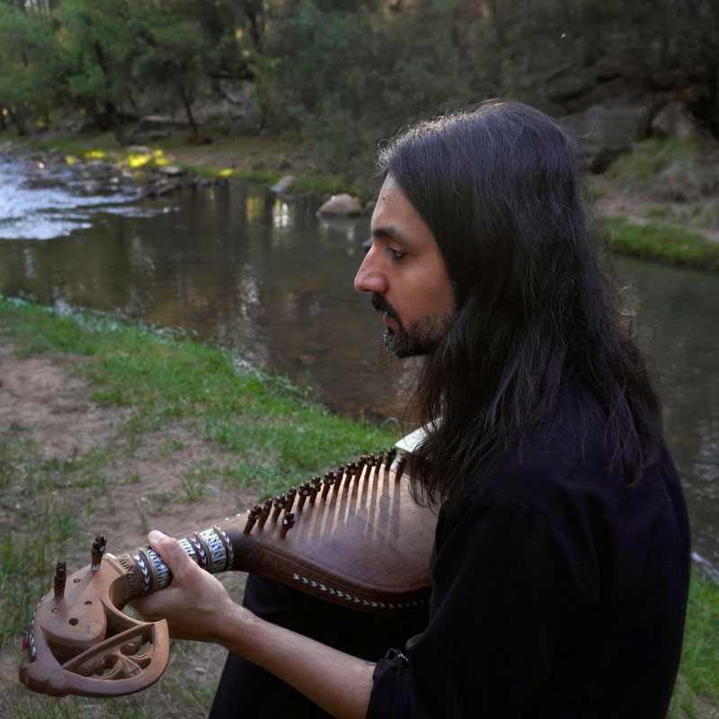 Qais Essar - Echoes of the Unseen . A photo of the artist playing the rabab by a river.