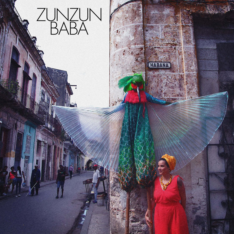 The single cover for Zunzun Baba by Rita Donte features a street scene in Havana, Cuba. A performer on stilts is dressed in a colorful costume resembling a hummingbird, with iridescent wings spread wide, a green-scaled bodysuit, and a feathered mask. Below, a woman in a flowing red dress and bold yellow accessories leans against the wall. The textured, weathered buildings and the "Habana" street sign anchor the image in a rich, cultural setting. The typography is minimal.