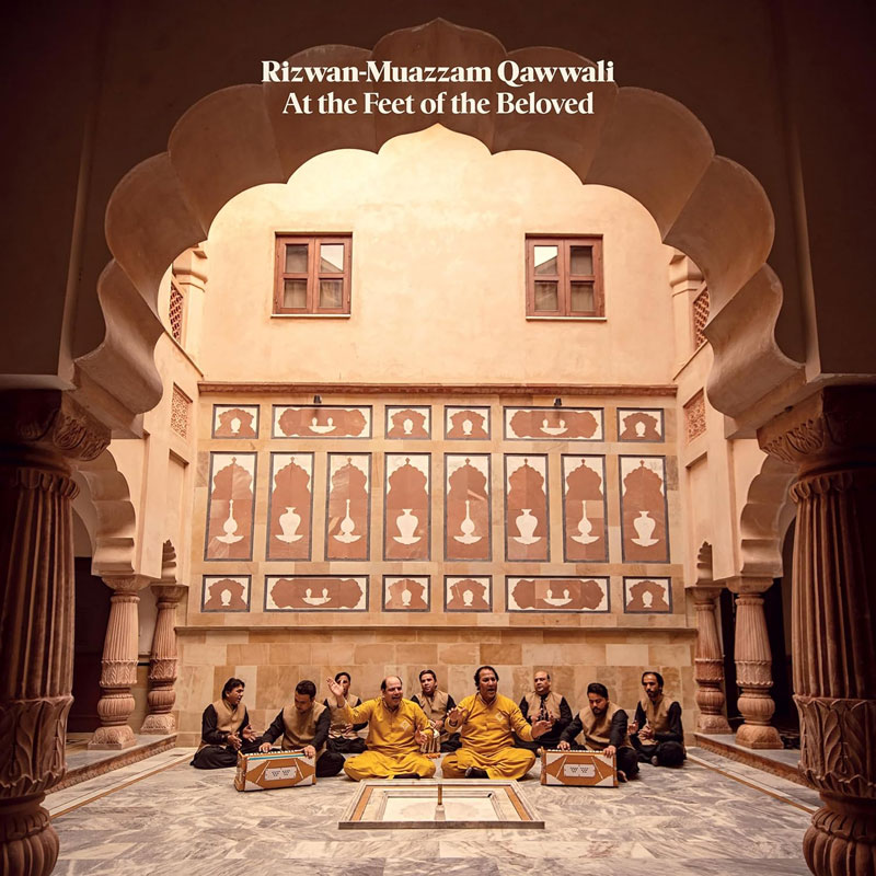 The album cover for At the Feet of the Beloved by Rizwan-Muazzam Qawwali features a symmetrical courtyard framed by ornate arches. The group, dressed in yellow and black traditional attire, is seated in a circle on a marble floor, centered beneath soft natural light. Behind them, a patterned wall of silhouettes and motifs adds depth.