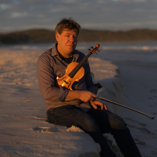 Robert Zielinski sitting outdoors, holding a violin, with a beautiful landscape in the background.