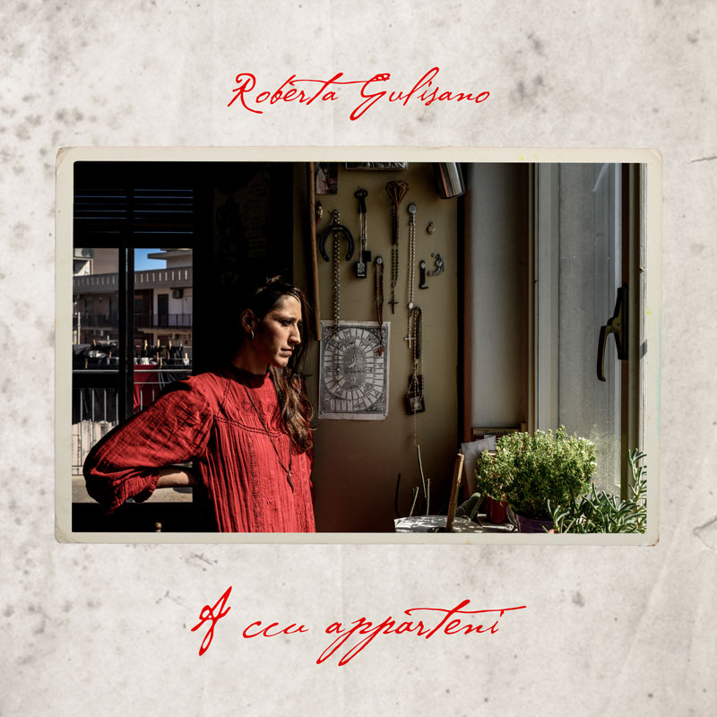The album cover for A Ccu Apparteni by Roberta Gulisano features a contemplative woman in a red dress standing indoors, gazing out a sunlit window. The warm-toned photo is bordered like a vintage snapshot, with delicate handwriting in red for the artist’s name and album title on a textured, parchment-like background.