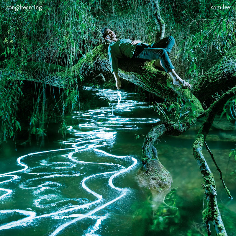 Sam Lee - Songdreaming cover artwork. the artist resting over a tree branch over a creek in a lush green forest.