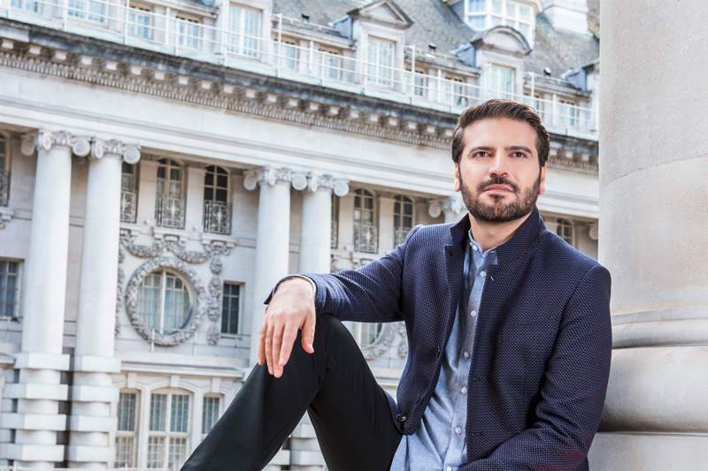 Sami Yusuf sitting leaning against a wall. An architecturally ornate building is behind him.