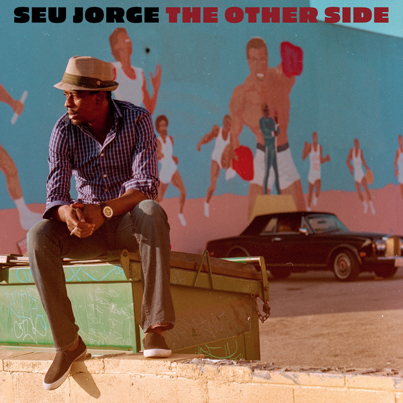 Seu Jorge sits alone on a weathered green dumpster beneath a mural of athletes and boxers, while a vintage black car drifts through the sunlit background.
