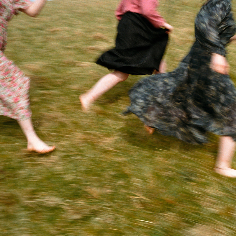 The cover features the bottom half of three wearing dresses and a skirt running across a grass field.