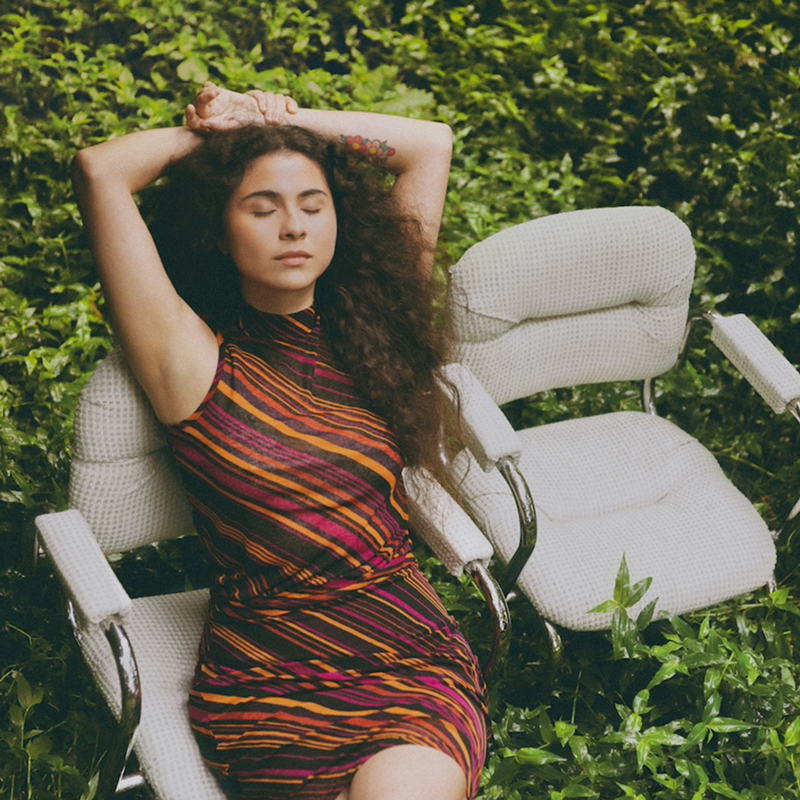 The cover of Vendrán Suaves Lluvias shows Silvana Estrada seated among lush green plants, eyes closed, arms resting behind her head. She wears a striped dress in shades of orange, magenta, and black.