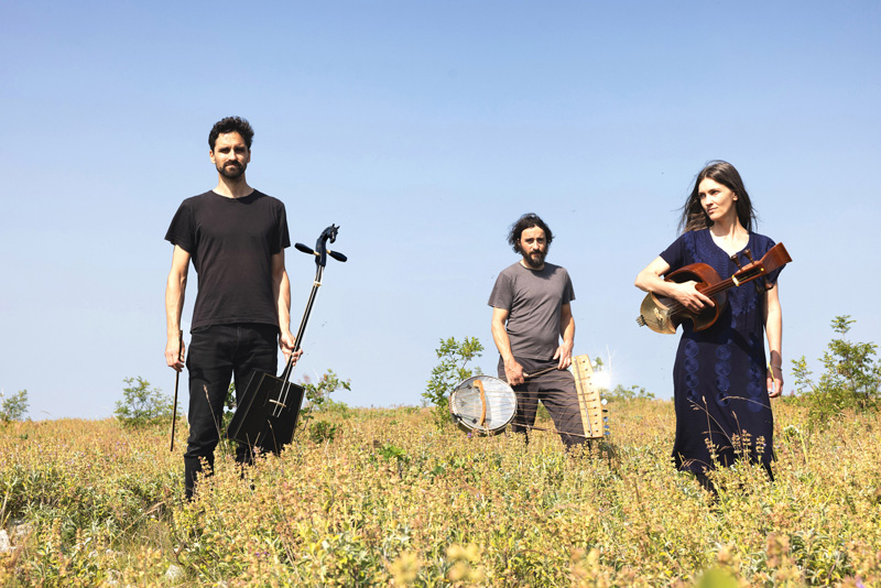 Širom - Photo by Nada Žgank . The trio outdoors in a field holding musical instruments.