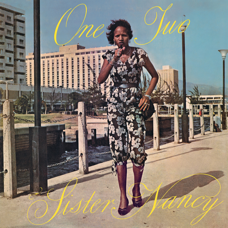 The album cover for One Two by Sister Nancy features the artist confidently walking toward the camera, microphone in hand, against a sunlit urban backdrop. Dressed in a patterned jumpsuit and heels. The scene is set by the waterfront with a hotel and mountains in the distance.