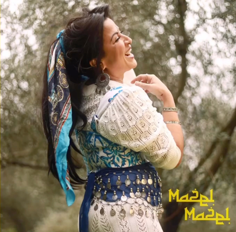 Syna Awel Mazel Mazel artwork. A color phoo of Syna smiling, wearing traditional clothes in a forest.